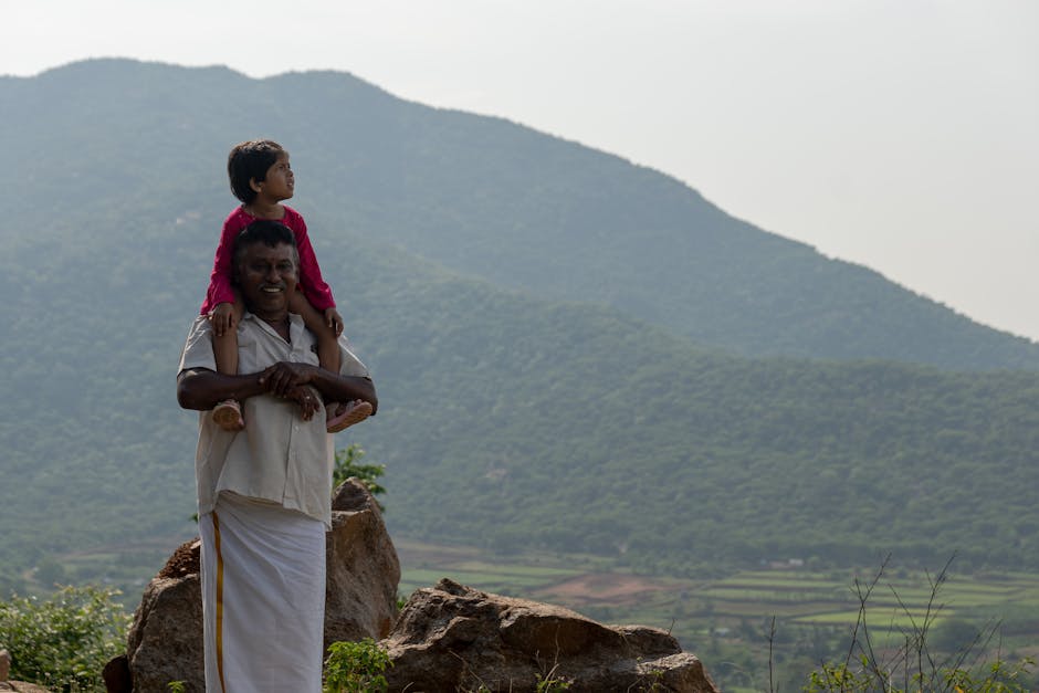 A joyful grandfather carrying his granddaughter on shoulders amidst a picturesque mountain backdrop.