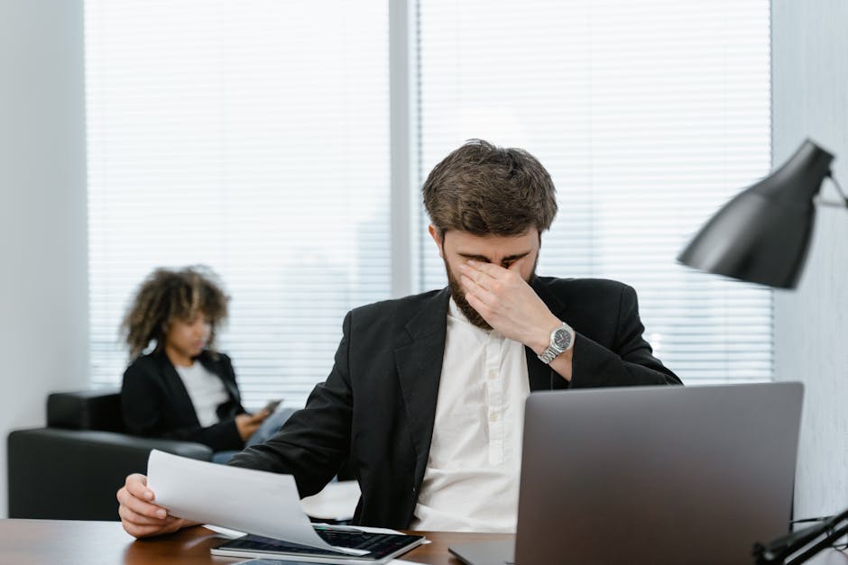 Businessman in an office covering his eyes while working on a laptop, feeling stressed.