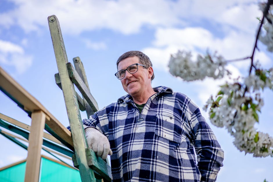 Smiling middle-aged man with glasses poses outdoors beside a wooden ladder on a sunny day.