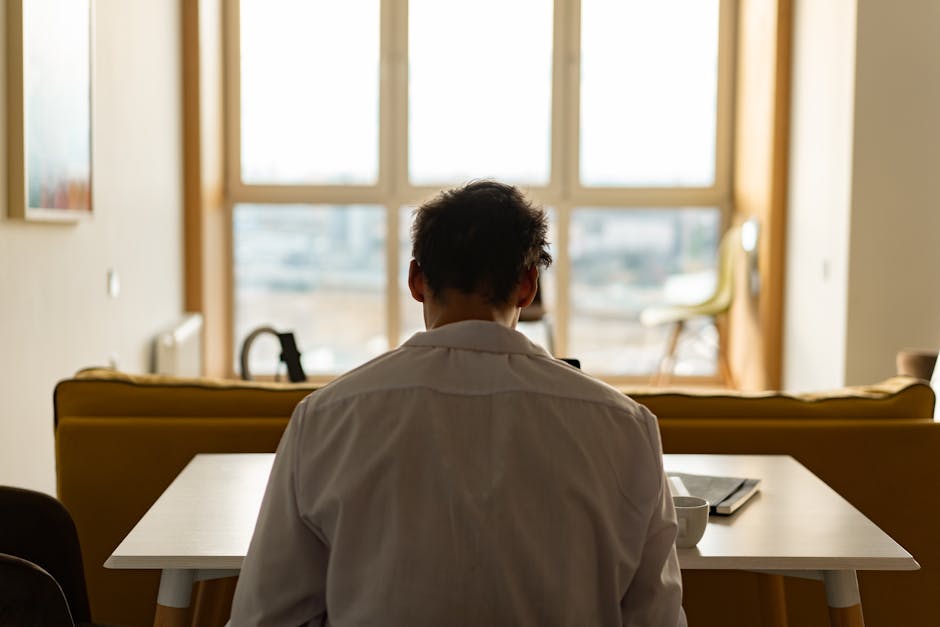 Back view of a man sitting at a desk, wearing a white shirt, with a window view.