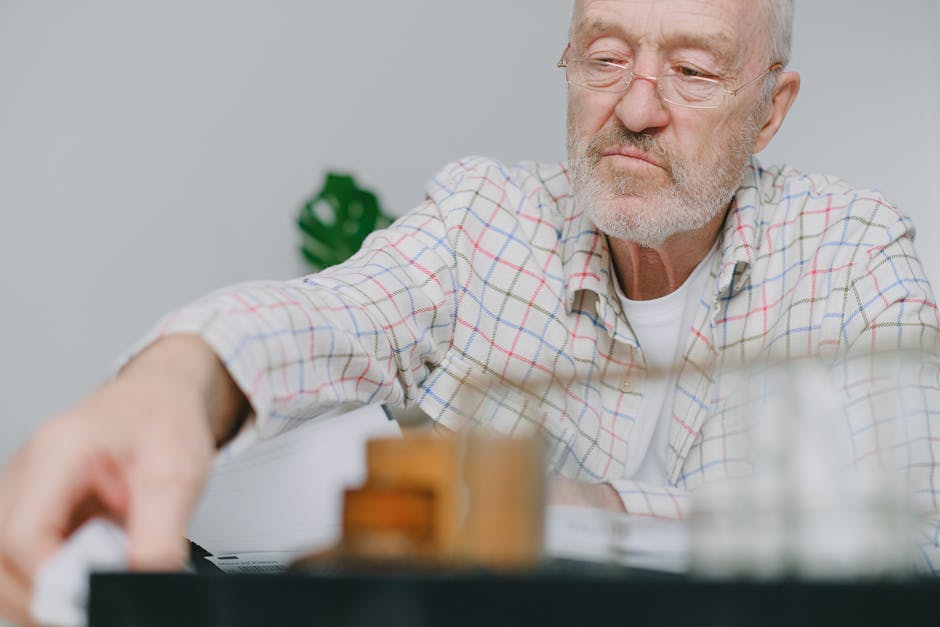 Elderly man wearing glasses, deep in thought indoors, blurred foreground.