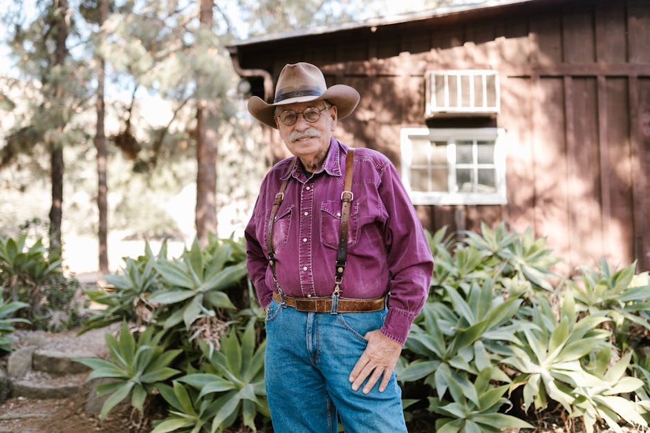 Senior man in cowboy hat smiling, standing outdoors by rustic building, surrounded by plants.