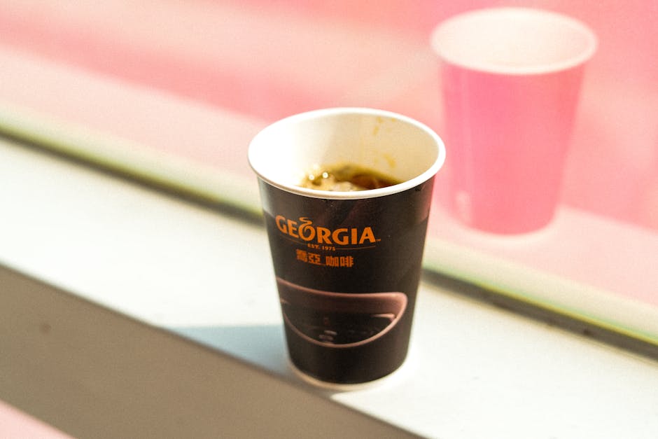 Disposable coffee cup from Georgia brand, resting on a sunlit windowsill with a blurred background.