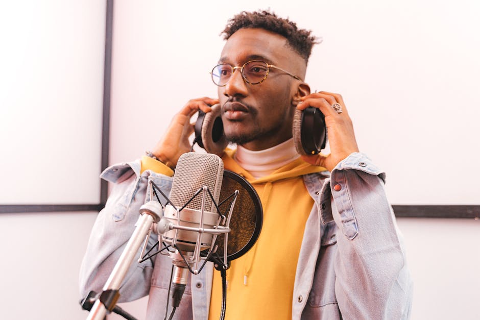 Confident young man singing in a studio with headphones, denim jacket, and microphone.