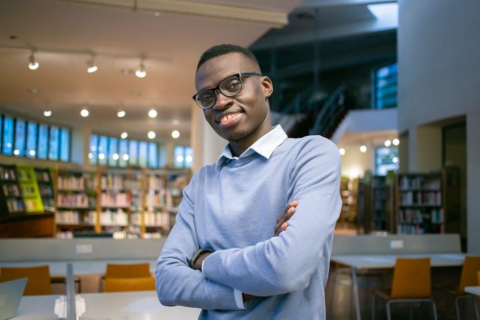 Positive African American student with crossed arms standing among tables and chairs in library