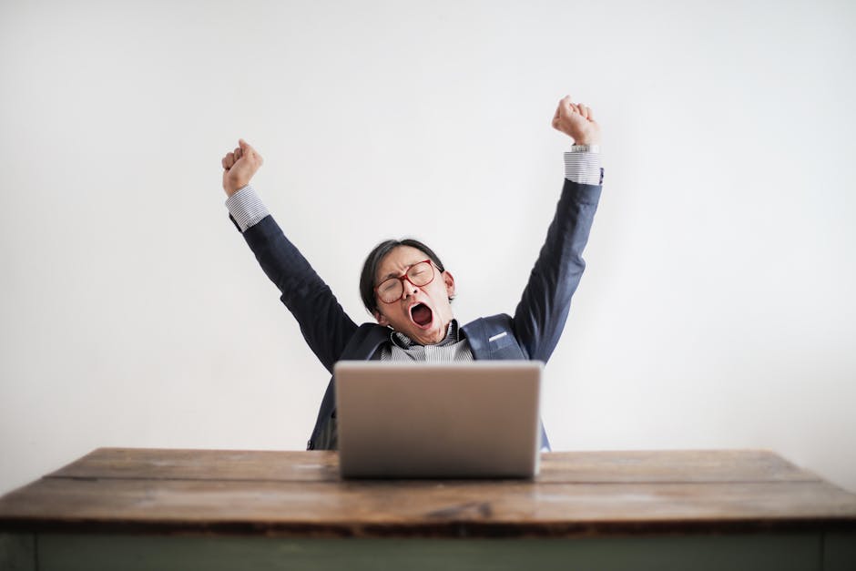 Exhausted man yawning at desk, working on a laptop in an office setting.
