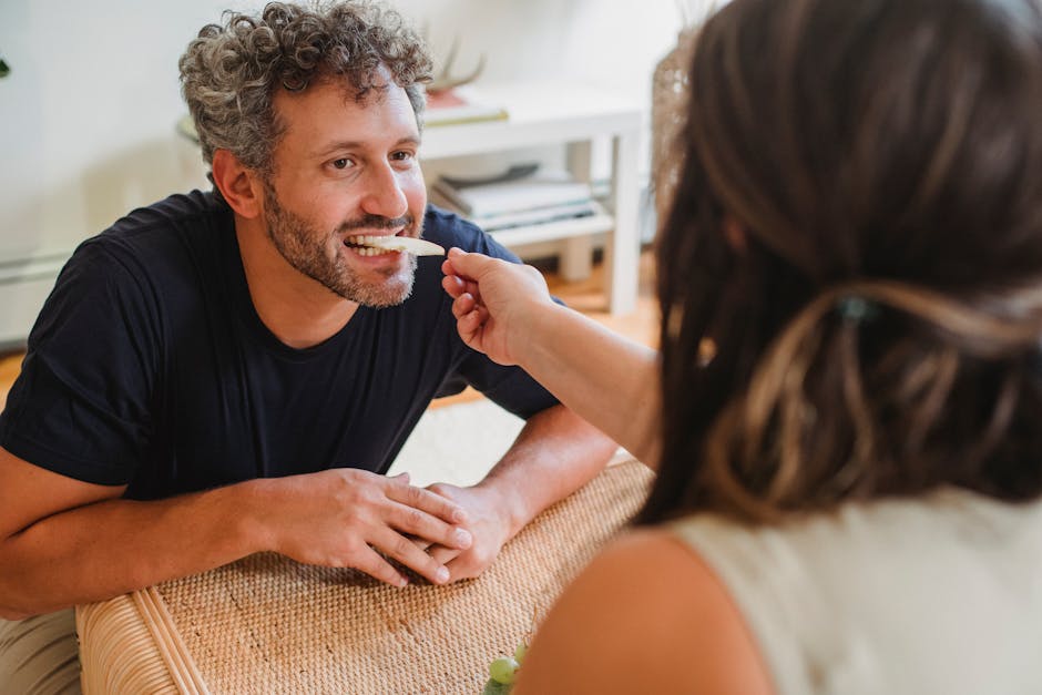 Cheerful bearded male with curly gray hair biting slice of apple from hand of wife