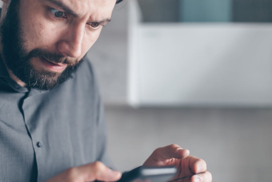 A bearded man in a gray shirt looks shocked while holding a smartphone indoors.
