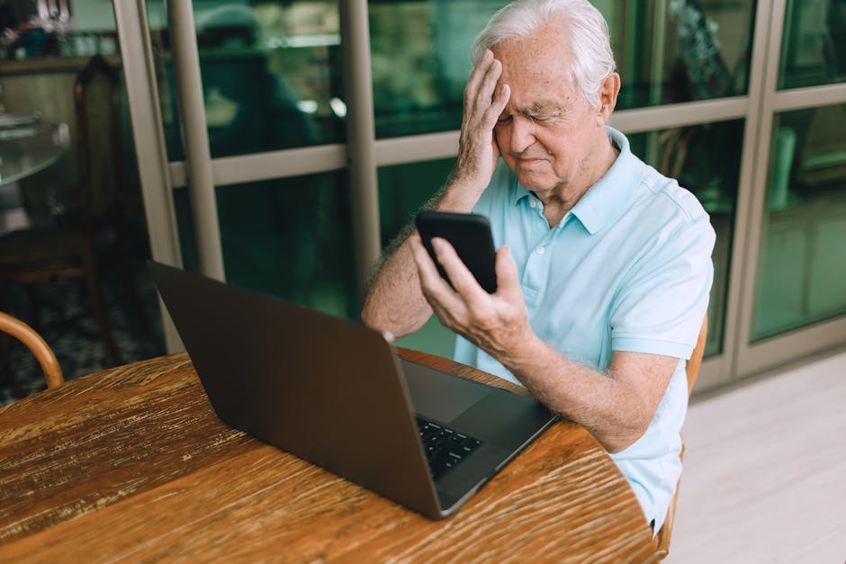 An elderly man in a blue shirt looks troubled while holding a smartphone and sitting at a table with a laptop.