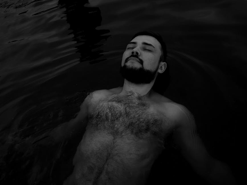 A serene black and white image of a shirtless man with a beard floating peacefully on water.