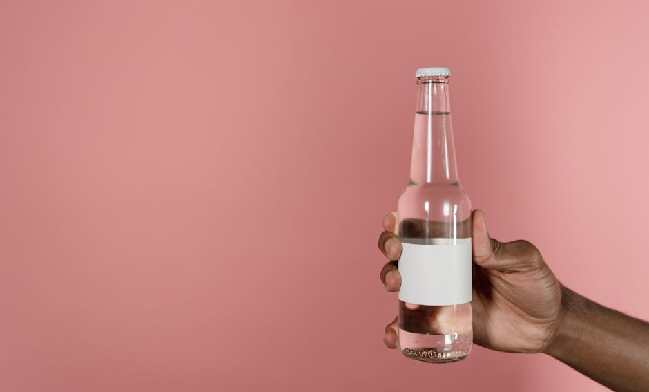 Close-up of a hand holding a clear glass bottle against a pink backdrop with space for design.