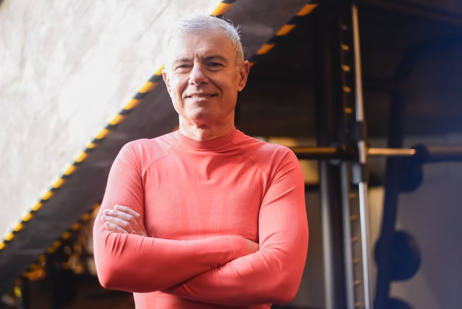 Elderly man wearing red shirt confidently poses with crossed arms at the gym.