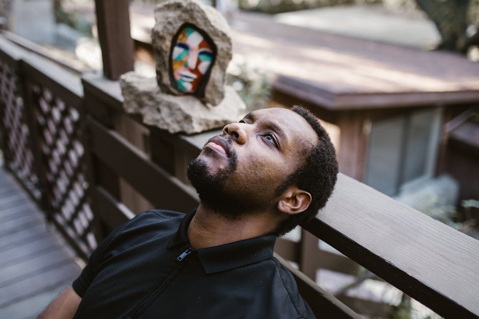 A man in thoughtful reflection sits outdoors against a wooden railing with abstract art behind.